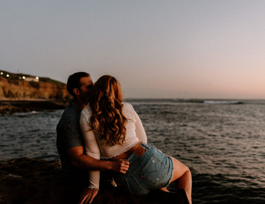 A photograph of a man and woman sitting near the ocean during their engagement session at Sunset Cliffs in San Diego, CA.