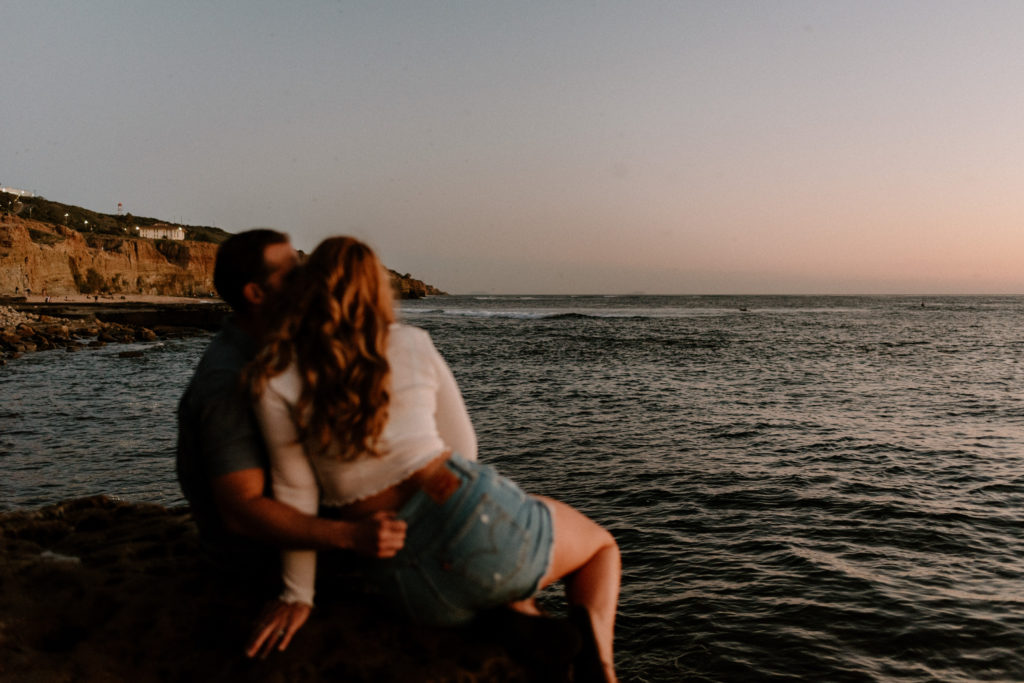 A photograph of a man and woman sitting near the ocean during their engagement session at Sunset Cliffs in San Diego, CA.