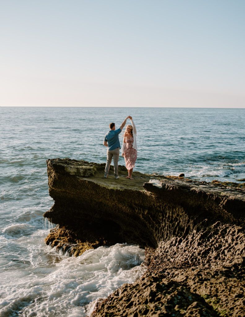 A photograph of a man and woman dancing near the ocean during their engagement session at Sunset Cliffs in San Diego, CA.